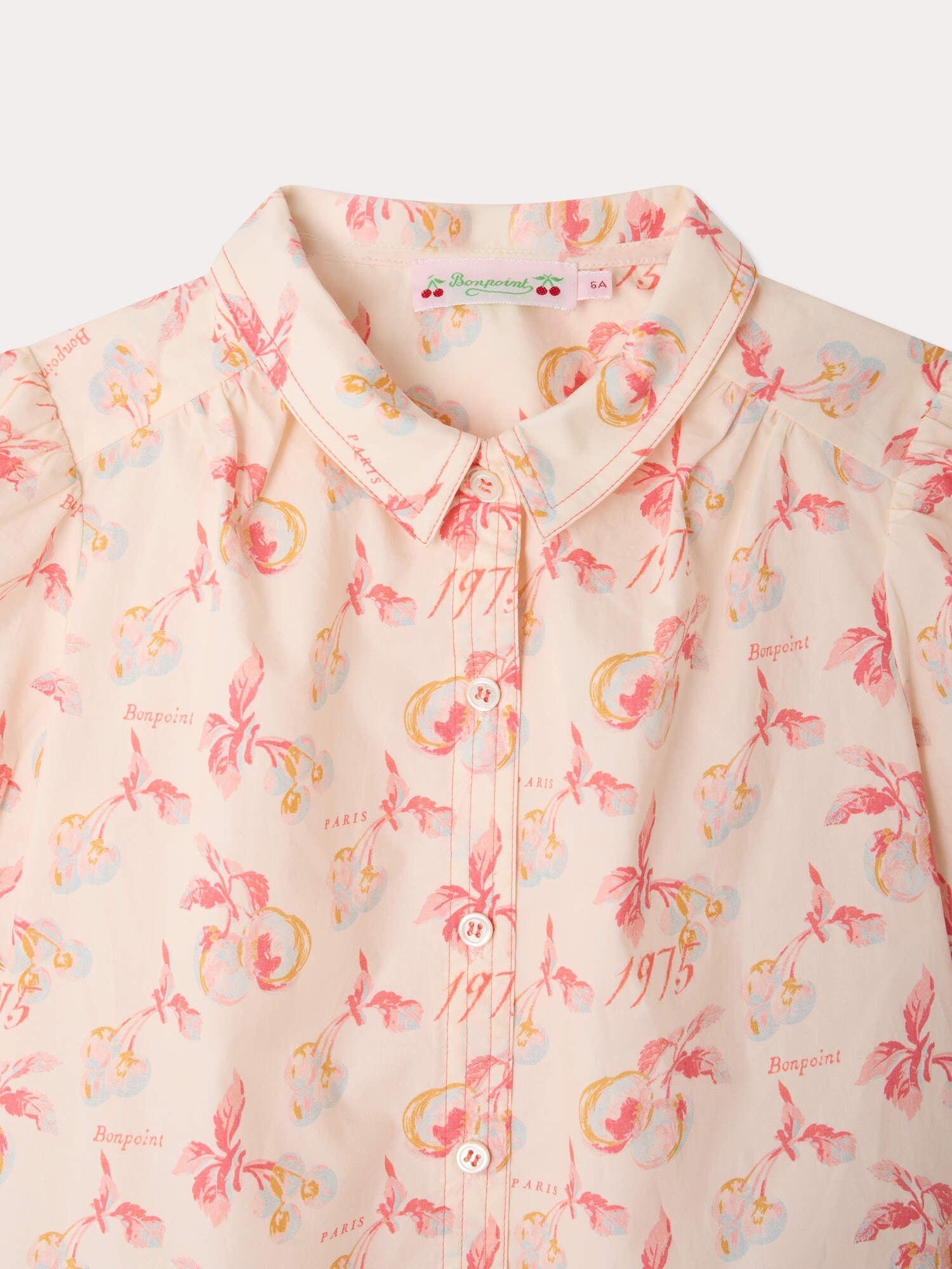 The Girls’ Mathilda Blouse by Bonpoint is a light pink collared blouse for girls, featuring a red and yellow floral print, a button-down front, and text such as “Bonpoint,” “PARIS,” and “1975” printed across the organic cotton fabric.