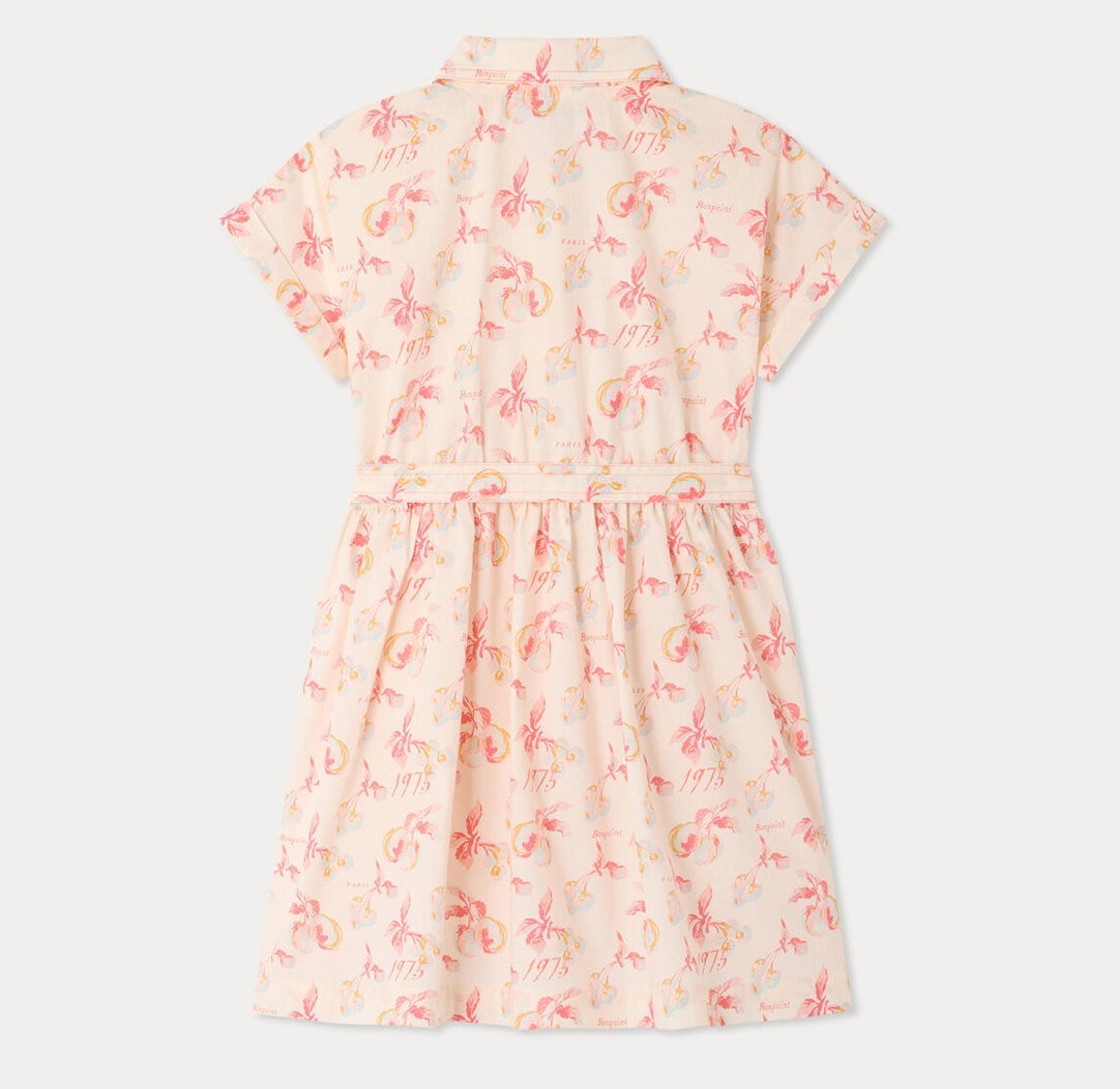 The Girls’ Gisele Dress by Bonpoint is a light pink shirt dress featuring short sleeves, a collar, and a button-down top with a belted waist. Made from organic cotton, it showcases an all-over tropical floral print with palm trees and “1975” in muted pink and orange tones.