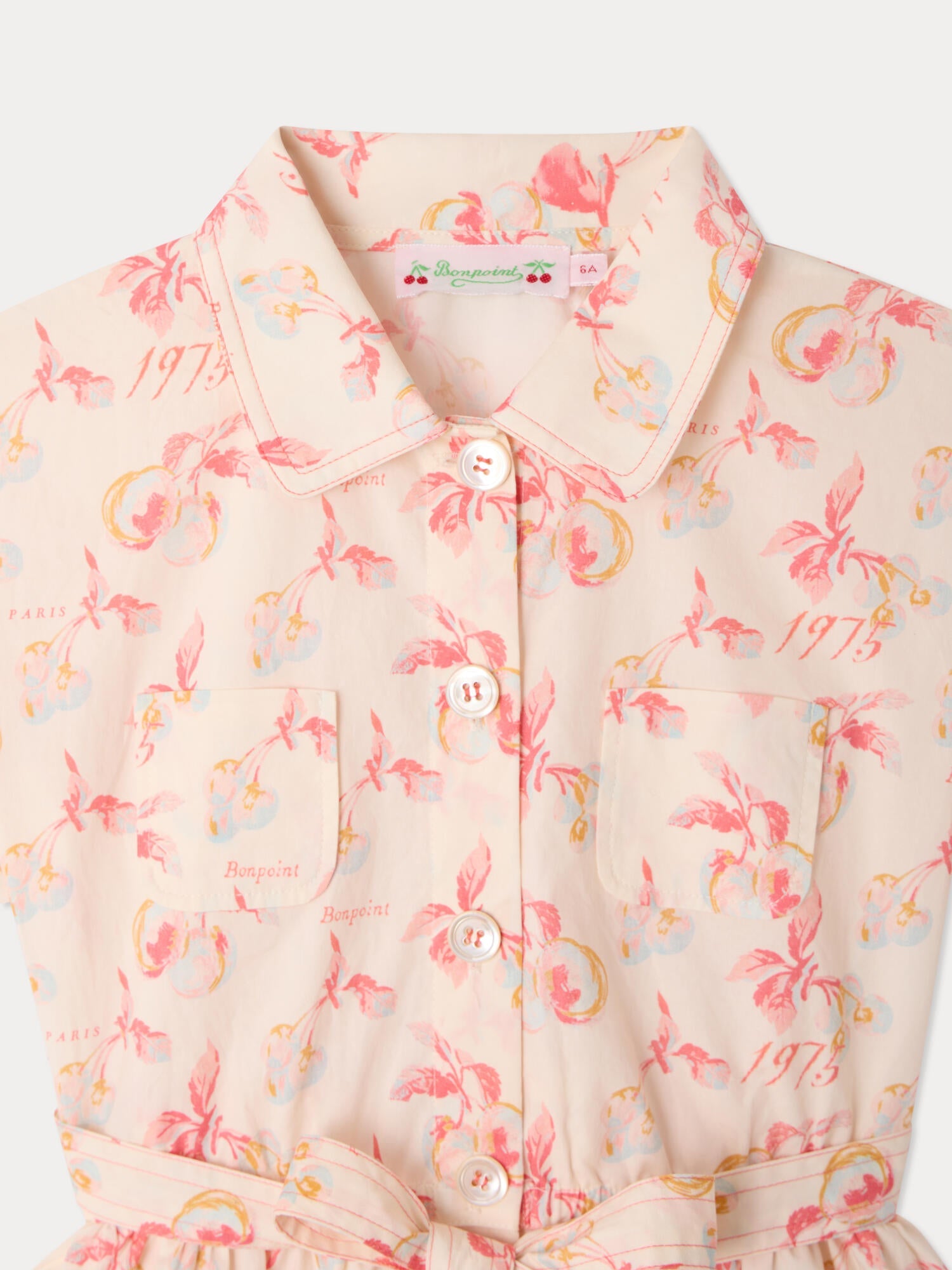 Close-up of the Girls’ Gisele Dress by Bonpoint in light beige organic cotton, featuring a pink floral and fruit print, white buttons, two front pockets, and scattered “1975” and “Paris” text on the fabric.