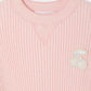 The Girls’ Melie Sweater by Bonpoint is a pink ribbed organic cotton sweater, adorned with a pastel cherry patch on the upper left chest and a triangular stitch detail below the collar. It features a subtle waffled texture, making it the perfect cozy girls’ sweater.