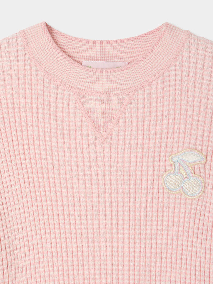 The Girls’ Melie Sweater by Bonpoint is a pink ribbed organic cotton sweater, adorned with a pastel cherry patch on the upper left chest and a triangular stitch detail below the collar. It features a subtle waffled texture, making it the perfect cozy girls’ sweater.