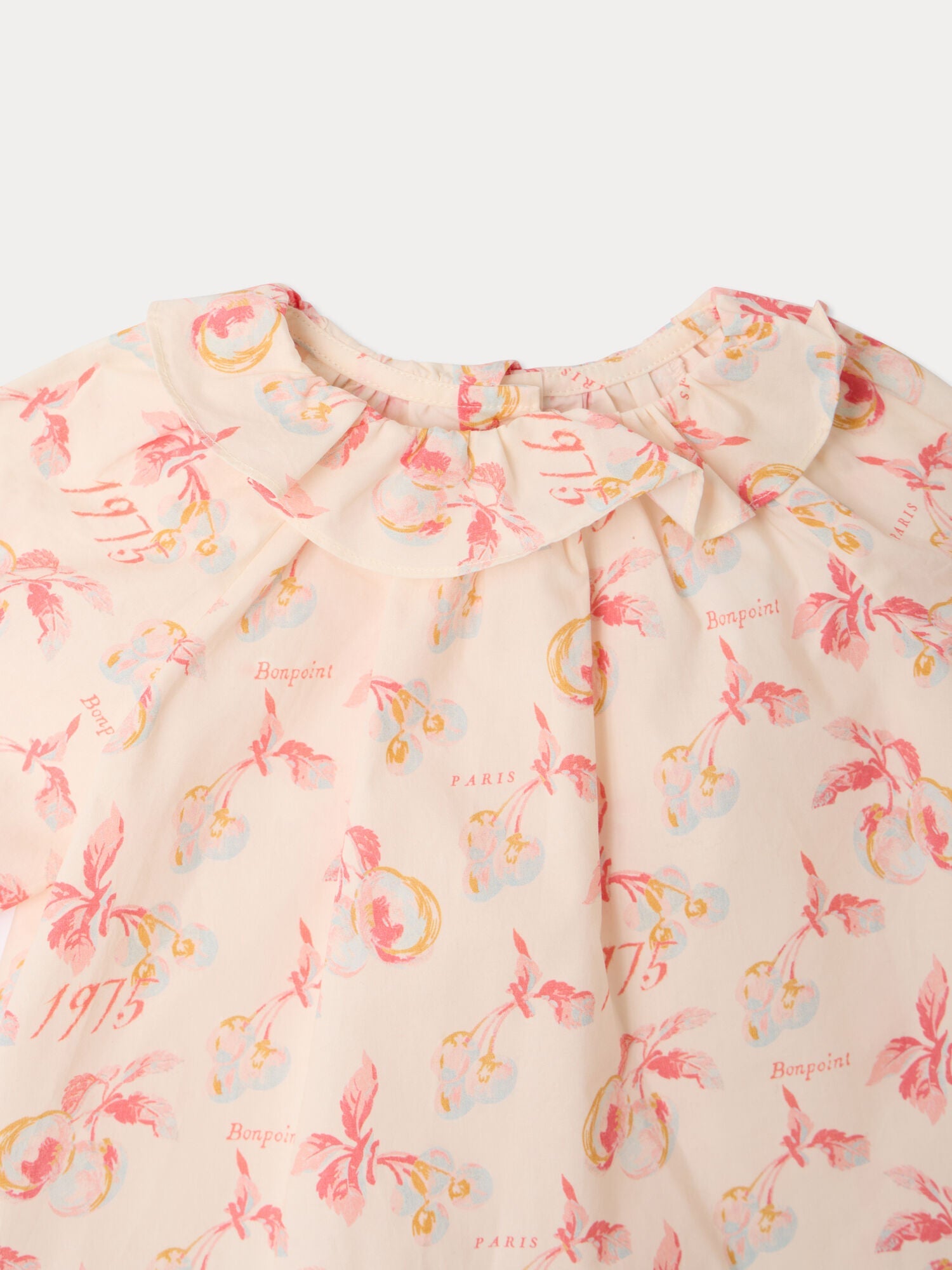 Close-up of the Bonpoint Baby Girls’ Dolci Blouse in light pink organic cotton, featuring a ruffled collar and cherry illustrations alongside “Bonpoint,” “1975,” and “Paris” text in soft pink and orange tones, elegantly finished with a delicate floral print.