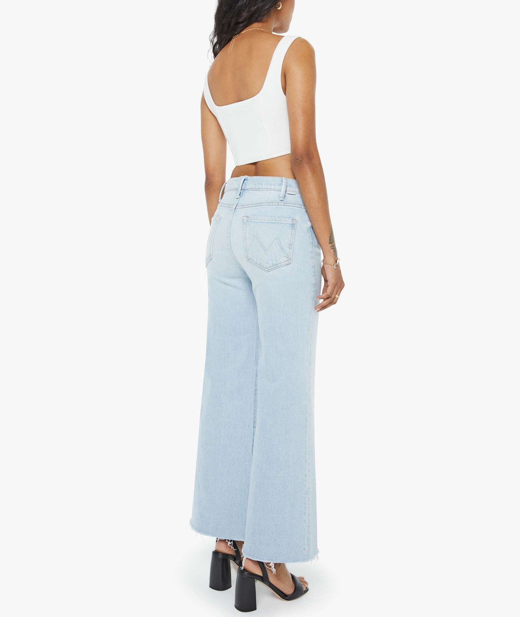 A woman stands with her back to the camera, wearing the Mother Petites Lil Hustler Roller Fray high-rise jeans in light blue, paired with a white sleeveless crop top and black heeled sandals, set against a plain white background.