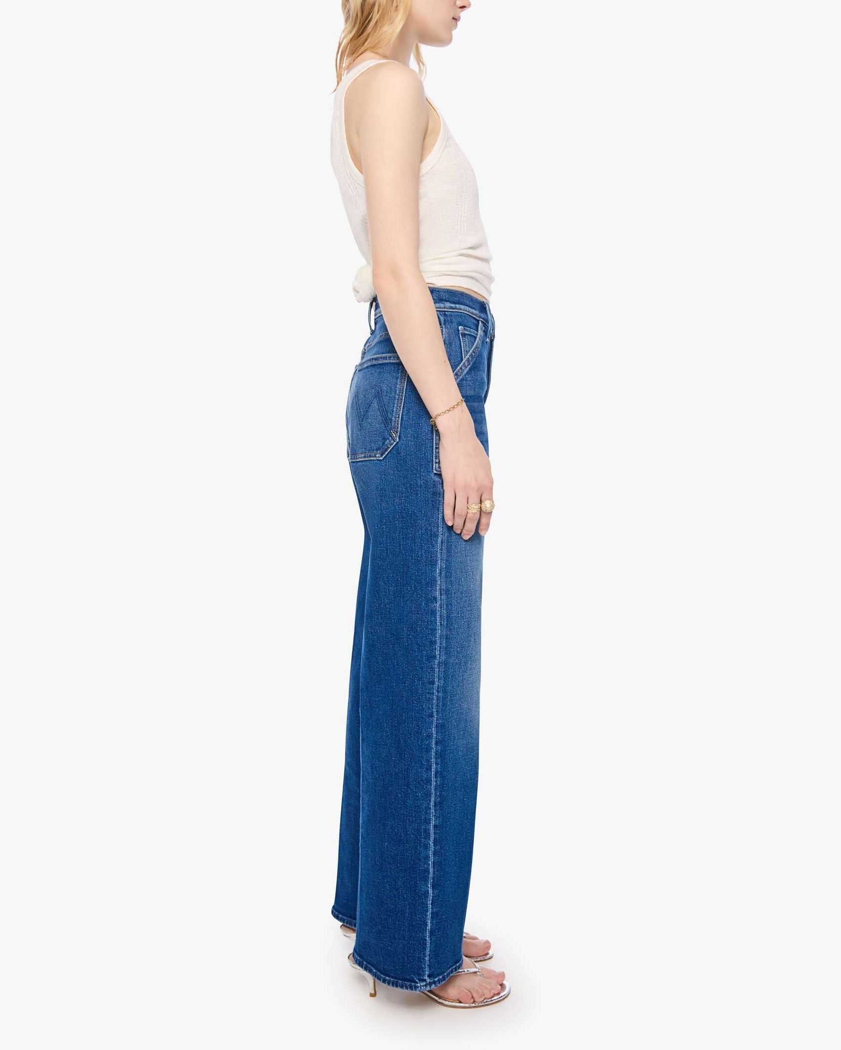 A woman stands in profile against a plain white background wearing the Mother Petites Lil Patch Maven Sneak: high-waisted, wide-leg blue jeans made from recycled stretch denim, paired with a white sleeveless top.