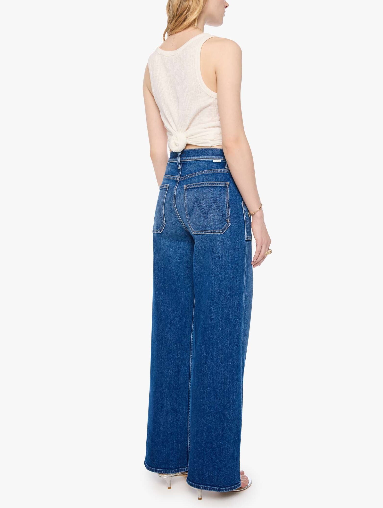 A woman stands facing away, wearing the Mother Petites Lil Patch Maven Sneak high-rise wide leg jeans crafted from recycled stretch denim with back pockets and a white sleeveless top tied at the back.