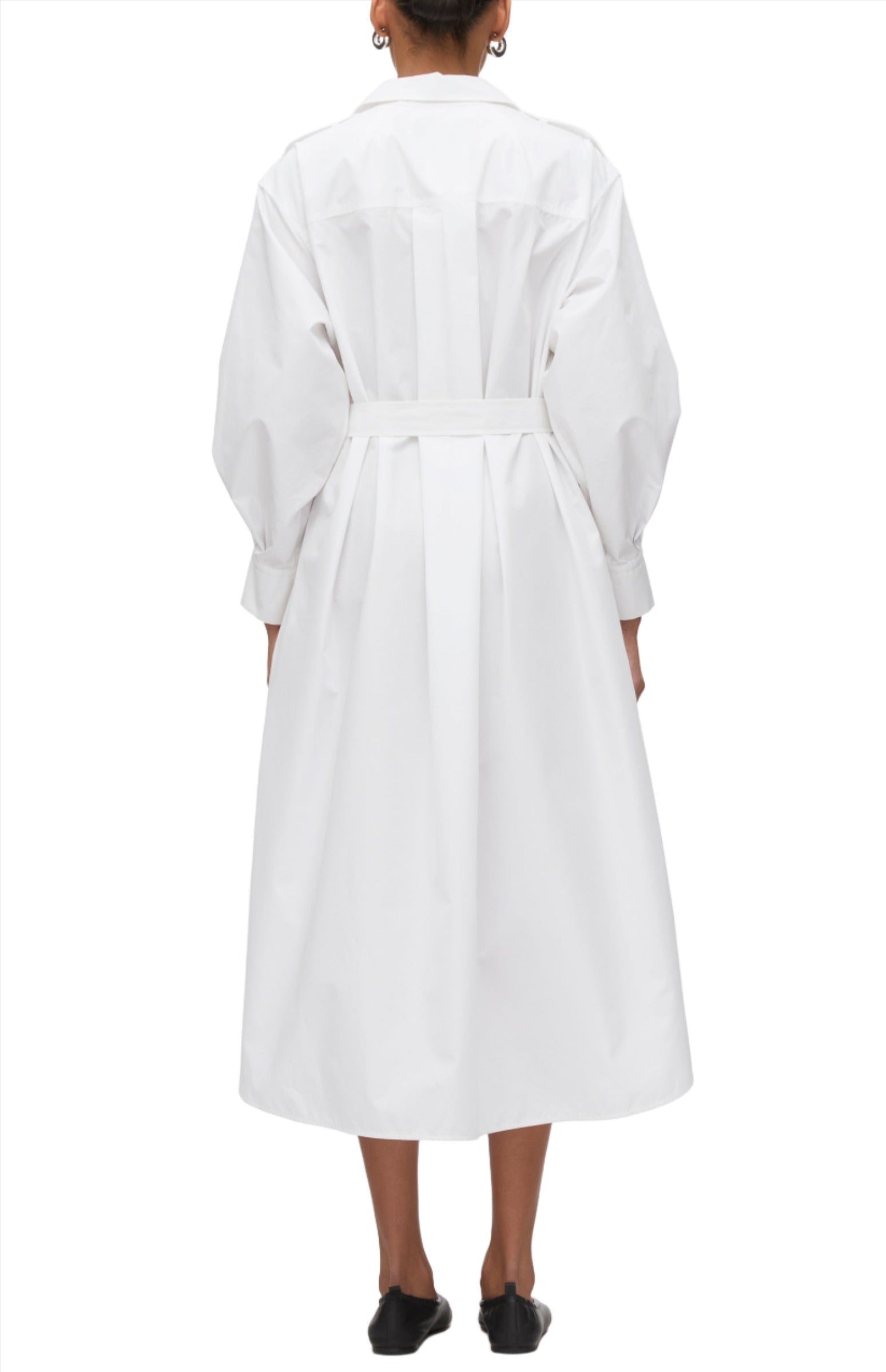 A person stands with their back to the camera, dressed in a 3.1 Phillip Lim Poplin Utility Midi Shirt Dress, which is a long-sleeved, ankle-length white coat cinched with a belt.