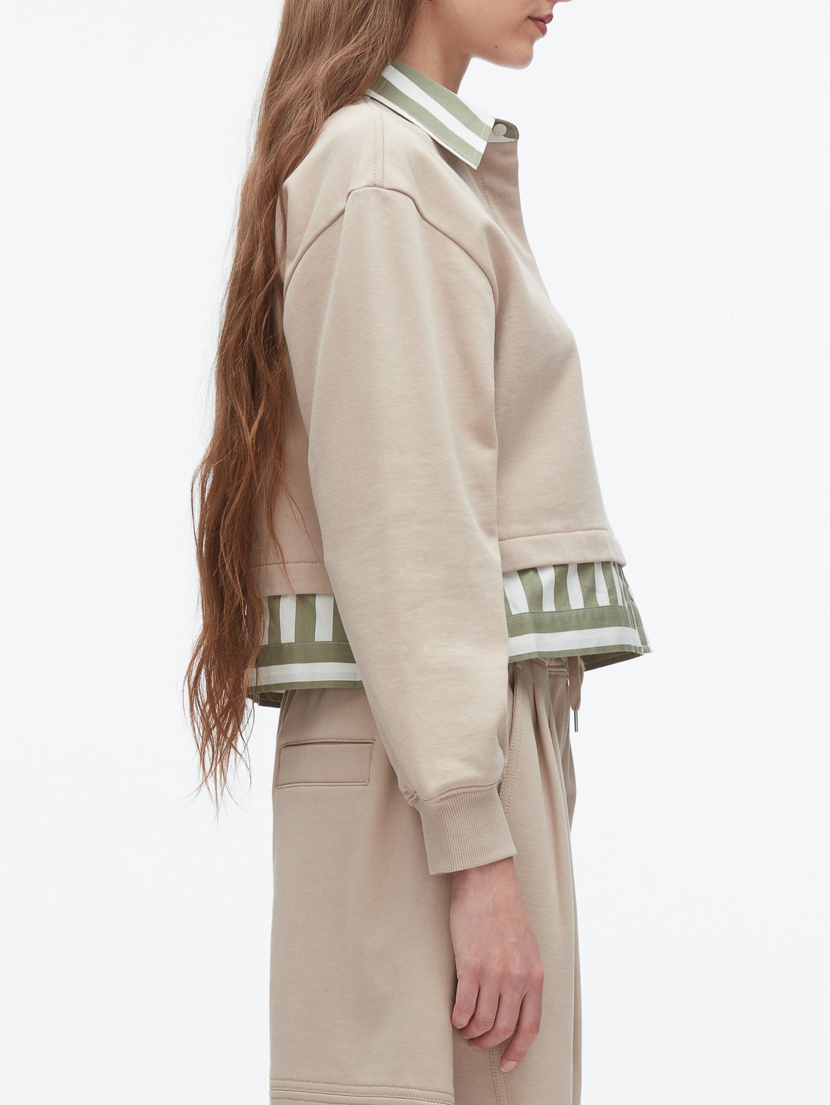 A woman stands in profile wearing the 3.1 Phillip Lim Cropped Polo with Stripe Combo Top—a beige long-sleeve layer with a green-and-white striped polo detail underneath, perfect for effortless layering.