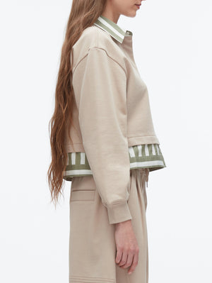 A woman stands in profile wearing the 3.1 Phillip Lim Cropped Polo with Stripe Combo Top—a beige long-sleeve layer with a green-and-white striped polo detail underneath, perfect for effortless layering.