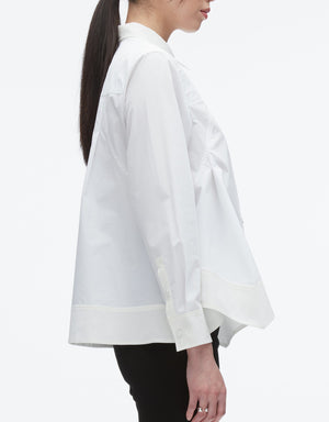 A woman in profile wears the 3.1 Phillip Lim Flounce Hem Poplin Shirt—a loose-fitting, long-sleeve white top with an asymmetric hem and cuffed sleeves—paired with black pants for a modern take on a classic look.