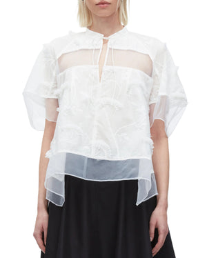 A woman wears the 3.1 Phillip Lim Embroidery Voile Top with Organza Combo—featuring sheer white embroidery, short sleeves, a layered hem, and keyhole neckline—paired with a black skirt.