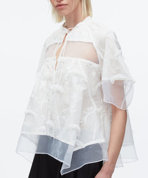 A person wears the 3.1 Phillip Lim Embroidery Voile Top with Organza Combo, a sheer white blouse featuring intricate embroidery, short sleeves, and a tie neckline.