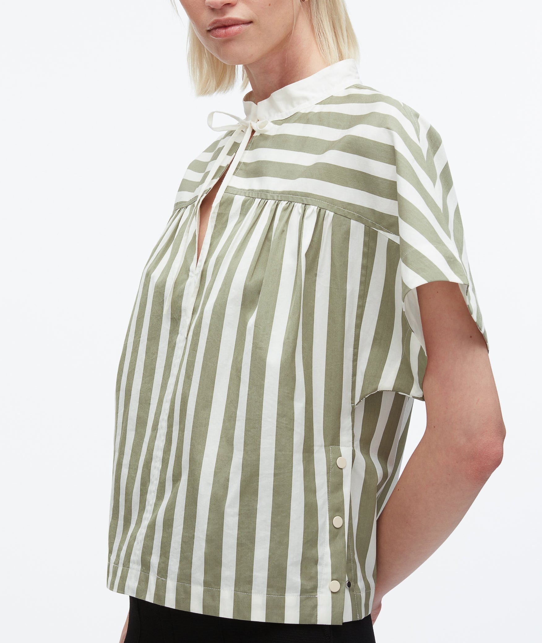 A person wearing the 3.1 Phillip Lim Striped Open Neck Top with Ties, a loose, short-sleeved green and white cotton poplin blouse featuring a tie neckline and side button details.