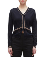 A woman wears the 3.1 Phillip Lim Chiffon Hem Boxy Cardigan, a varsity-inspired black V-neck with beige trim and long sleeves, featuring a sheer chiffon waist panel for a modern twist, paired with black pants.