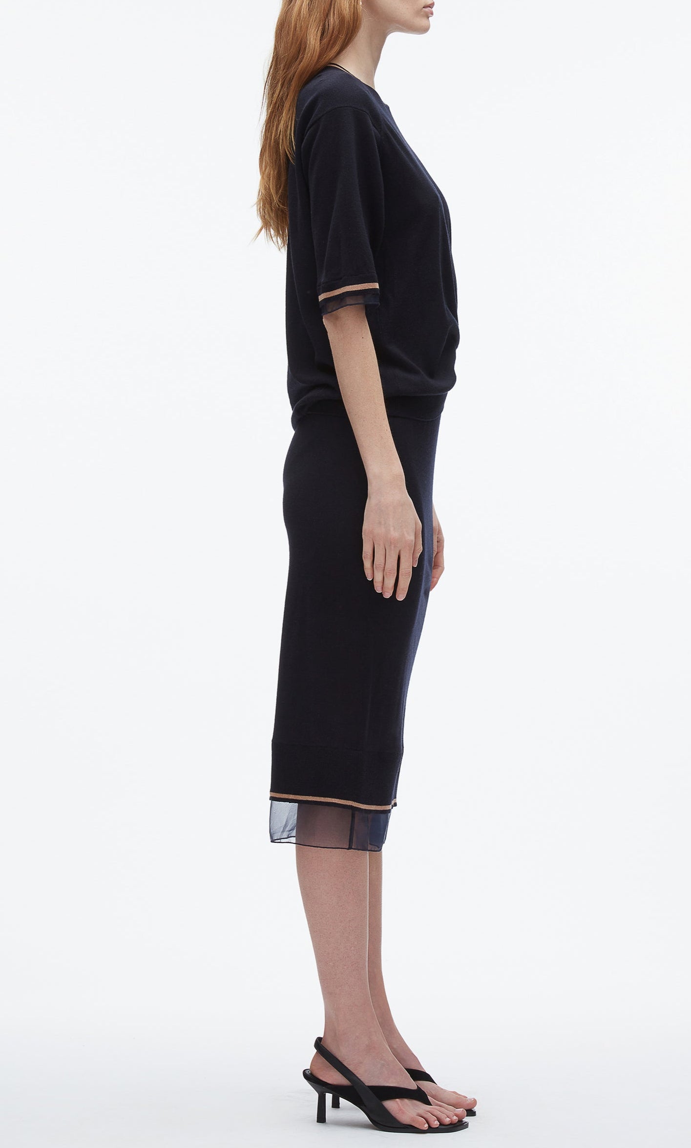 A woman stands in profile wearing the 3.1 Phillip Lim French Tuck Knit Dress with Organza, a black body-skimming silhouette featuring sheer trim, paired with black heeled sandals.