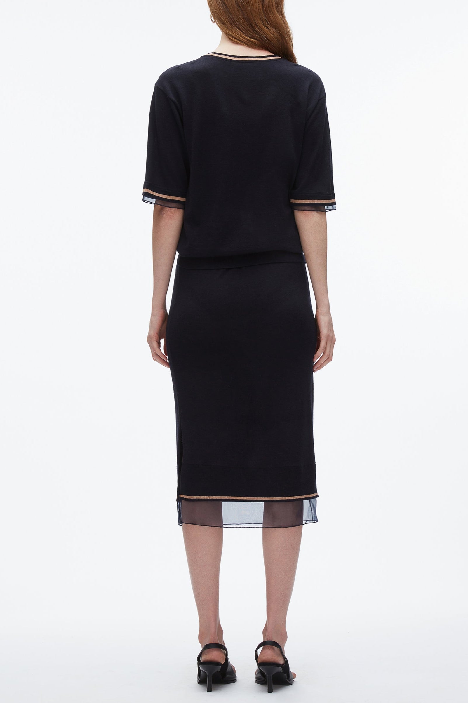 A woman stands with her back to the camera, wearing the 3.1 Phillip Lim French Tuck Knit Dress with Organza, styled with black heeled sandals.
