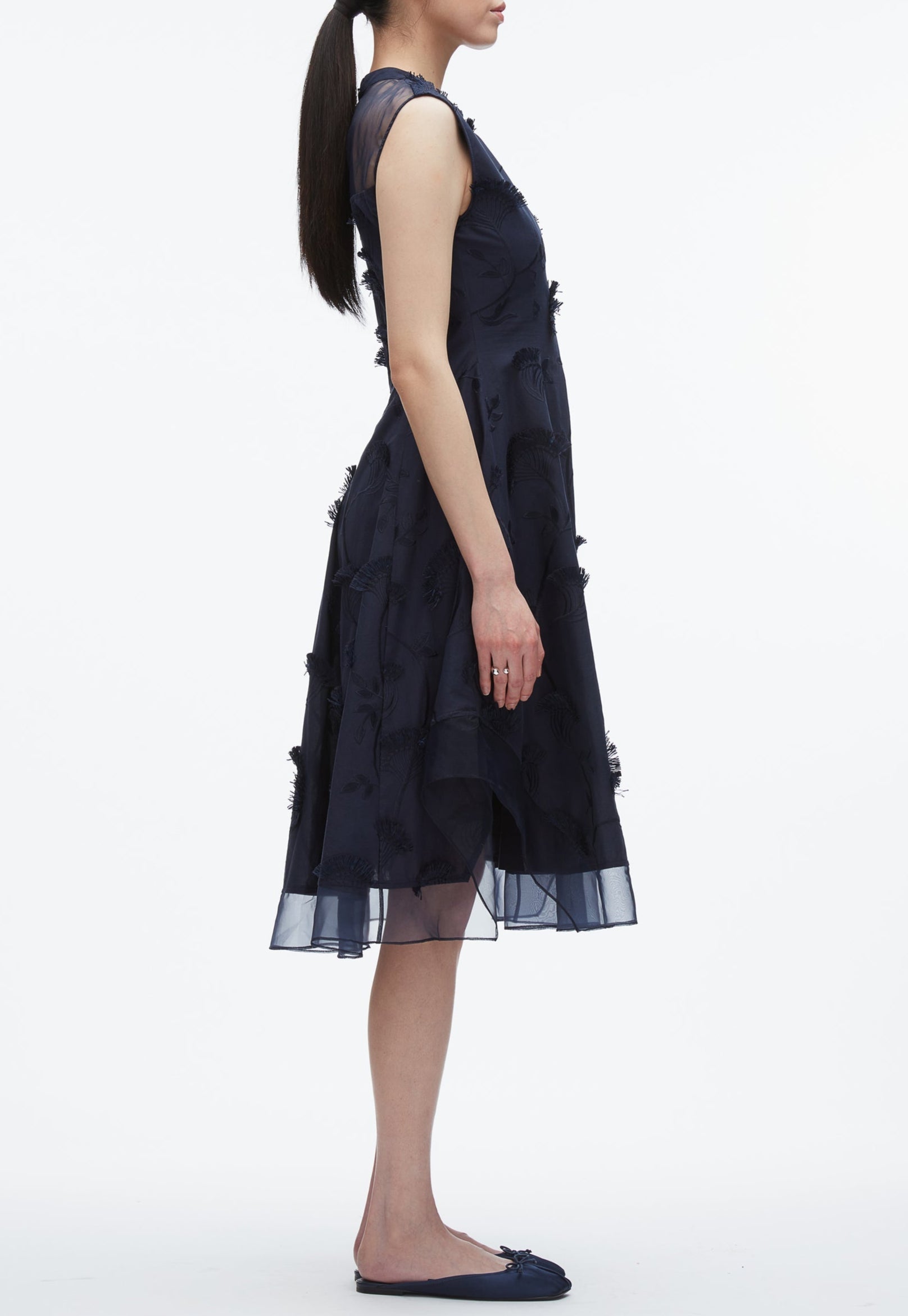 A woman in profile wears the 3.1 Phillip Lim Fil Coupé Midi Dress—navy blue, sleeveless cotton voile with embroidered floral accents—paired with navy flats and a ponytail, posing against a white background.