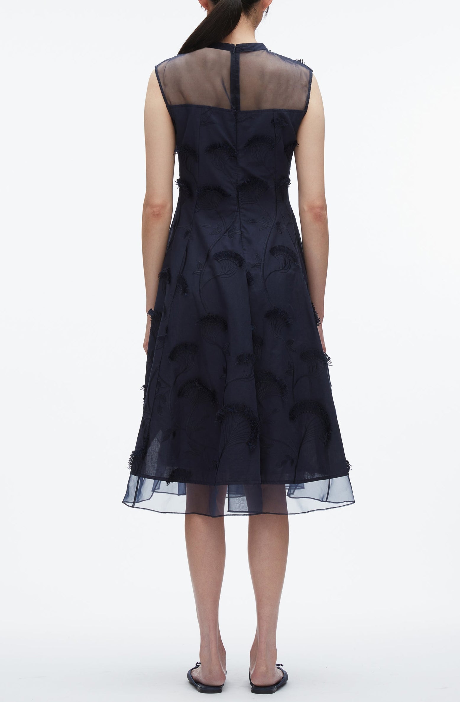 A woman faces away, wearing the 3.1 Phillip Lim Fil Coupé Midi Dress in navy cotton voile with a sheer upper back and embroidered florals, paired with flat sandals.