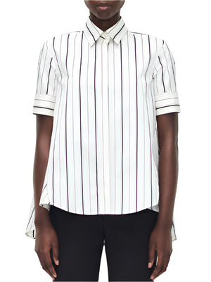 A person wearing the Adam Lippes Trapeze Stripe Shirt—a short-sleeve, ivory and black striped button-up—and black pants, photographed from the front.