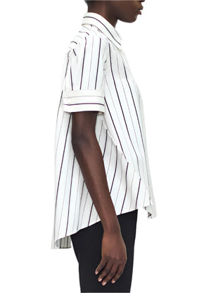 A person in profile wears the Adam Lippes Trapeze Stripe Shirt, featuring loose-fitting short sleeves and ivory and black stripes.