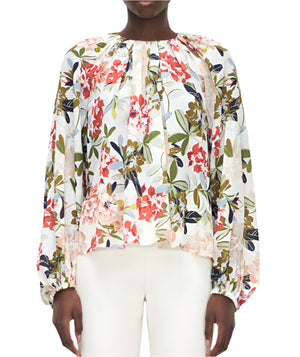 A person wears the Adam Lippes Lottie Top, a long-sleeve crepe de chine blouse with an ivory floral motif, paired with white pants and shown from the front.