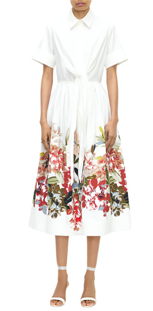 A woman wears the Adam Lippes Attacher Dress, a white collared short-sleeve midi dress with a waist sash and colorful floral print on the skirt, paired with white sandals.