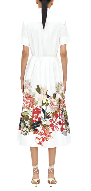 A person faces away, wearing the Adam Lippes Attacher Dress—an Italian cotton poplin collared style with floral print on the lower half, a waist sash, and strappy white heels.