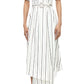 A person wears the Adam Lippes Nansi Stripe Dress—a white, short-sleeve, belted midi dress in striped poplin with vertical black stripes and an asymmetrical hem—with black strappy heels against a white background.