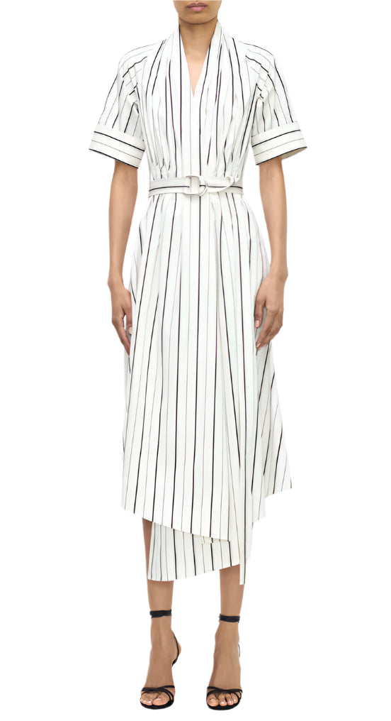 A person wears the Adam Lippes Nansi Stripe Dress—a white, short-sleeve, belted midi dress in striped poplin with vertical black stripes and an asymmetrical hem—with black strappy heels against a white background.