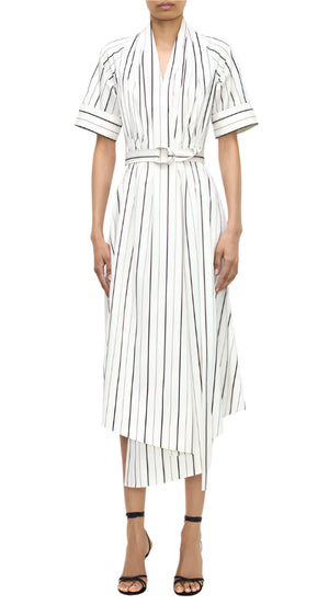 A person wears the Adam Lippes Nansi Stripe Dress—a white, short-sleeve, belted midi dress in striped poplin with vertical black stripes and an asymmetrical hem—with black strappy heels against a white background.