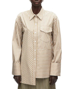 A person wears the 3.1 Phillip Lim Asymmetrical Stripe Trapeze Shirt, featuring beige and white mixed stripes, an asymmetrical chest pocket, and a relaxed silhouette.