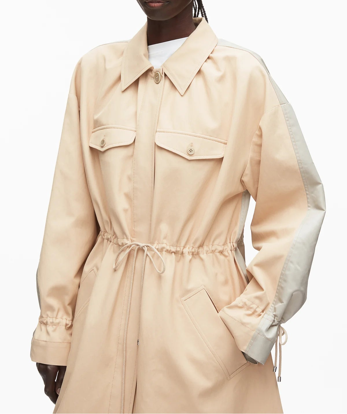 A person wearing the 3.1 Phillip Lim Blouson Trench Coat, a beige cotton-viscose utility jacket featuring buttoned chest pockets, side pockets, drawstring waist, and contrasting light sleeve panels for a modern touch.