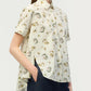 A woman wears the Adam Lippes Charlie Top, a short-sleeved, white button-up featuring a delicate floral print and classic collared design, paired with dark blue jeans. She stands slightly turned to the side with one hand in her pocket.