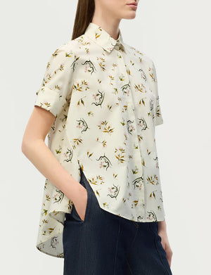 A woman wears the Adam Lippes Charlie Top, a short-sleeved, white button-up featuring a delicate floral print and classic collared design, paired with dark blue jeans. She stands slightly turned to the side with one hand in her pocket.