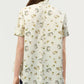 A person is shown from the back against a plain background, wearing the Adam Lippes Charlie Top— a light-colored collared blouse adorned with a subtle floral and line drawing pattern.