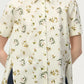 A person wearing the Adam Lippes Charlie Top—a short-sleeved, cream-colored blouse with a collared silhouette and floral and animal print—stands against a plain background.