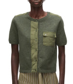 A person wearing the 3.1 Phillip Lim Merino Surplus Combo Top—a fitted, olive green cardigan with vintage military style, short sleeves, button front closure, chest pocket, and mixed fabric panels.