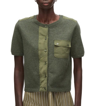 A person wearing the 3.1 Phillip Lim Merino Surplus Combo Top—a fitted, olive green cardigan with vintage military style, short sleeves, button front closure, chest pocket, and mixed fabric panels.