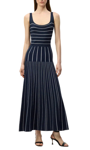 A woman wears the Adam Lippes Gemma Compact Jacquard Dress, a sleeveless navy blue fit and flare style featuring white vertical and horizontal stripes, paired with black heeled sandals.