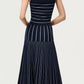 A woman stands with her back to the camera, wearing the Adam Lippes Gemma Compact Jacquard Dress—a sleeveless navy fit and flare style featuring white vertical and horizontal stripes—paired with black heeled sandals against a plain light background.