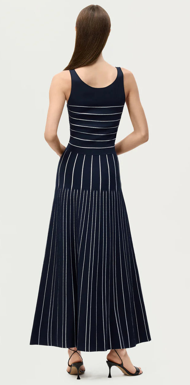 A woman stands with her back to the camera, wearing the Adam Lippes Gemma Compact Jacquard Dress—a sleeveless navy fit and flare style featuring white vertical and horizontal stripes—paired with black heeled sandals against a plain light background.
