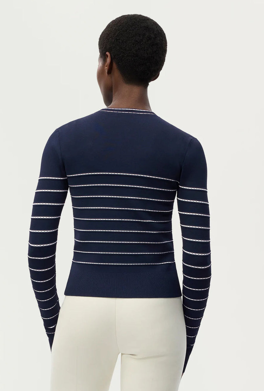A person is seen from behind, wearing the Adam Lippes Faye Cardigan—featuring navy and white horizontal stripes—with off-white pants, standing against a plain light background.