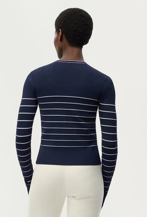 A person is seen from behind, wearing the Adam Lippes Faye Cardigan—featuring navy and white horizontal stripes—with off-white pants, standing against a plain light background.