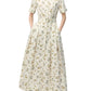 A woman wears the Leighton Dress by Adam Lippes, a white belted floral-print midi in Italian cotton poplin with short sleeves and button-down front, paired with cream low-heeled slingback shoes.