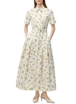 A woman wears the Leighton Dress by Adam Lippes, a white belted floral-print midi in Italian cotton poplin with short sleeves and button-down front, paired with cream low-heeled slingback shoes.