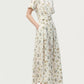 A woman wearing the Leighton Dress by Adam Lippes—a white, short-sleeve, collared maxi with a floral print and belted waist—pairs it with beige low-heeled shoes and stands with her hands in her pockets.