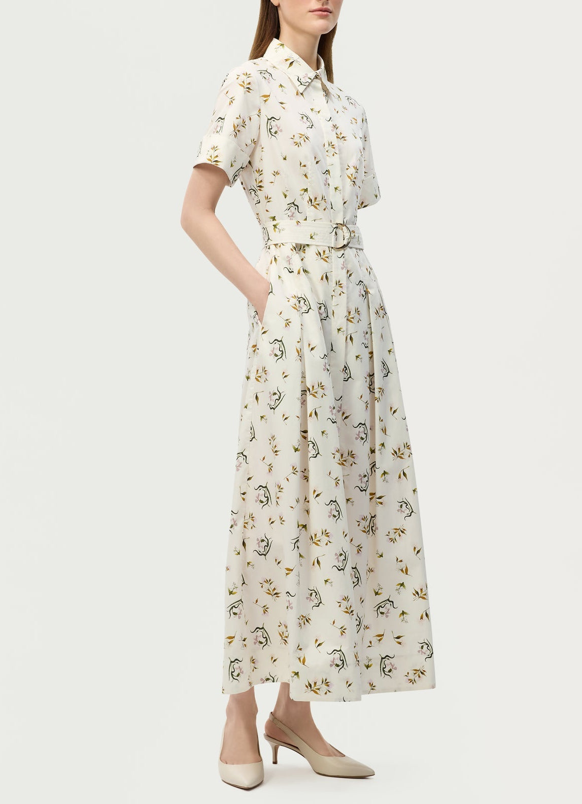A woman wearing the Leighton Dress by Adam Lippes—a white, short-sleeve, collared maxi with a floral print and belted waist—pairs it with beige low-heeled shoes and stands with her hands in her pockets.