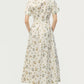 A woman faces away, wearing the Adam Lippes Leighton Dress—white Italian cotton poplin with a floral design—paired with beige heels for an elegant look.