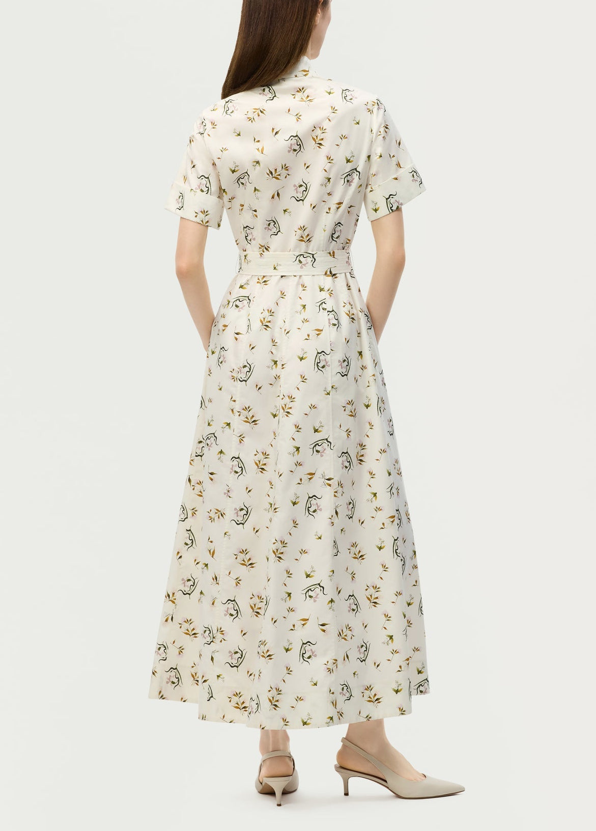 A woman faces away, wearing the Adam Lippes Leighton Dress—white Italian cotton poplin with a floral design—paired with beige heels for an elegant look.