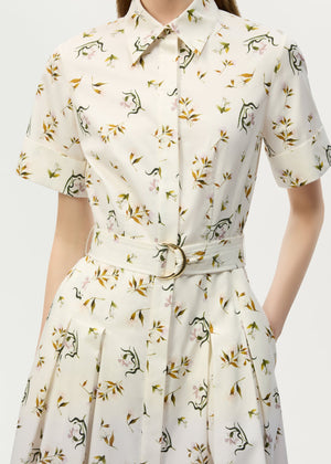 A woman wears the Leighton Dress by Adam Lippes, a short-sleeve, belted, collared button-up crafted from Italian cotton poplin with a playful dinosaur and leaf print on a light background.