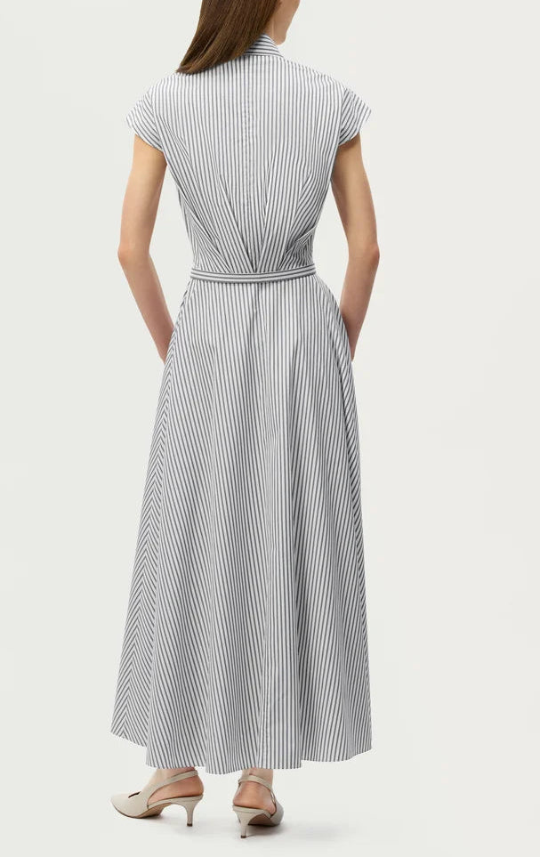 A person stands facing away, wearing the Adam Lippes Palmer Stripe Shirting Dress—a blue and white vertical striped, belted short-sleeve midi dress crafted from Italian cotton shirting—styled with cream low-heeled shoes, showcasing its long fluid silhouette.
